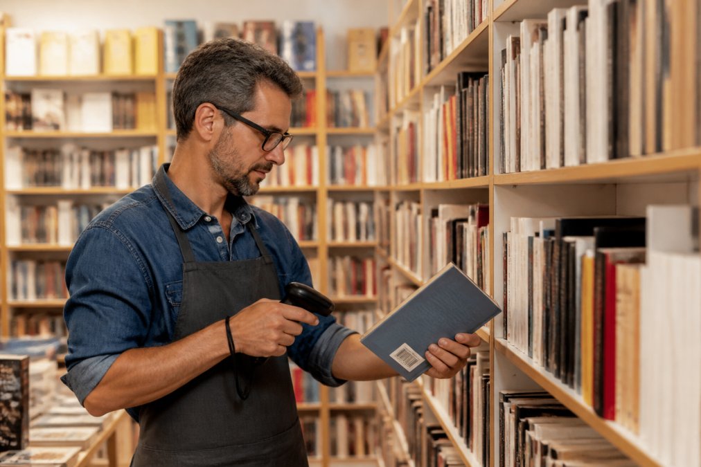 Libraio che scansiona il codice a barre di un libro con un lettore barcode portatile tra gli scaffali di una libreria indipendente ben illuminata, con scaffali di libri colorati sullo sfondo