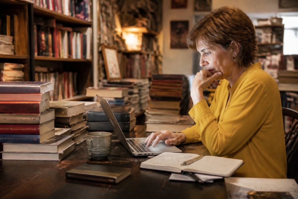 Independent bookstore owner, middle-aged woman, sitting at a wooden desk reviewing inventory data on a laptop screen, surrounded by stacks of books, handwritten notes and a cup of coffee, warm shop lighting in the background