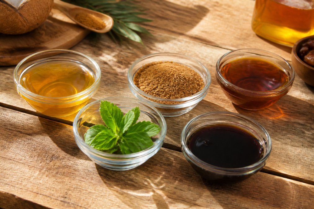 Verschiedene natürliche Süßungsmittel in Glasschüsseln nebeneinander arrangiert: Agavendicksaft, Kokosblütenzucker, Ahornsirup, Stevia-Blätter und Dattelsirup mit natürlichem Licht auf Holztisch