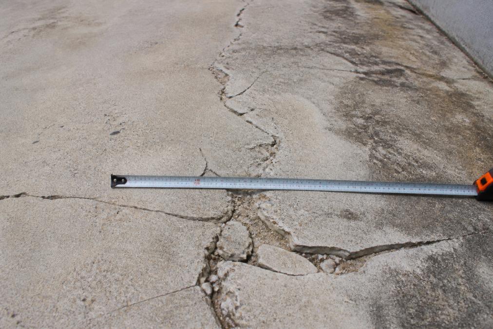 Close-up of a deteriorated concrete driveway surface showing deep cracks, discolouration stains, and spalling edges, with a metal measuring tape placed across the cracks for scale