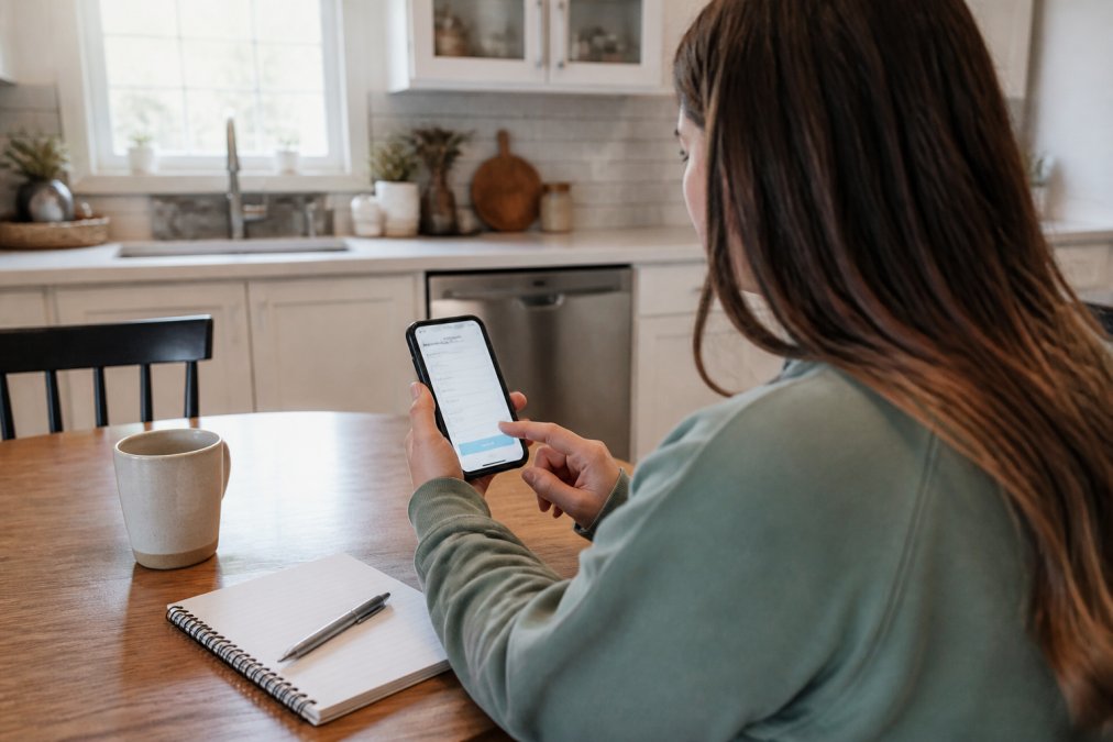 A homeowner sitting at a kitchen table with a smartphone, reviewing a cleaning quote form on the screen, with a notebook and coffee cup nearby
