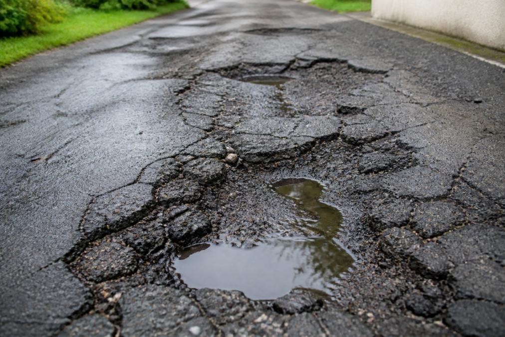 Close-up of severely cracked and potholed asphalt driveway showing rutting, surface deterioration, and water pooling in damaged areas