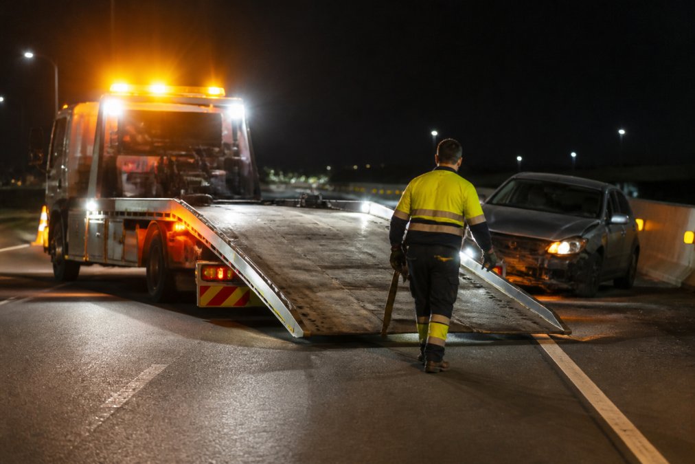Tow truck with flashing lights arriving at a nighttime accident scene, with recovery operator preparing equipment near a damaged vehicle
