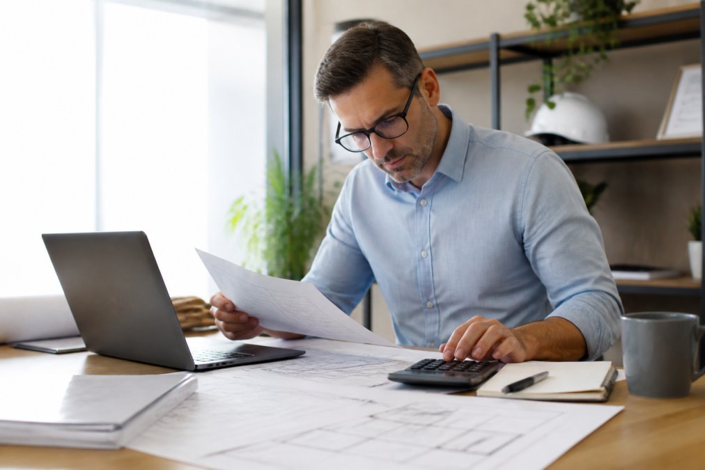Professional reviewing construction blueprints and financial documents at a desk with a laptop and calculator in modern office setting with natural lighting