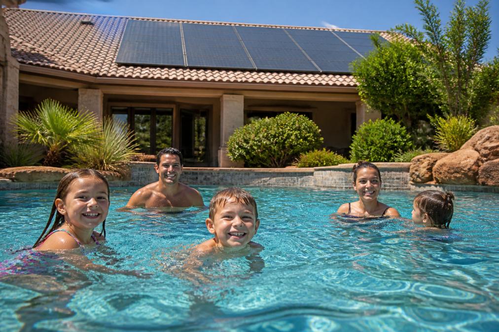Family enjoying a heated swimming pool outdoors on a sunny day with solar panels visible on the roof in the background, showing children and adults swimming while the sun reflects off the water