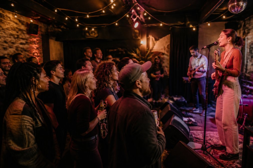 Diverse group of people enjoying a live concert performance in an intimate venue, audience members smiling and engaged with performers on stage, warm stage lighting creating vibrant atmosphere