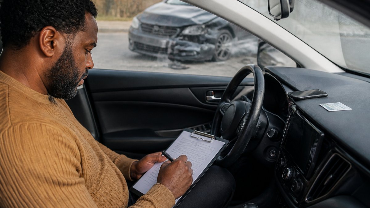 Driver sitting in car after accident, reviewing documents and taking notes on a clipboard with phone and insurance card visible on dashboard