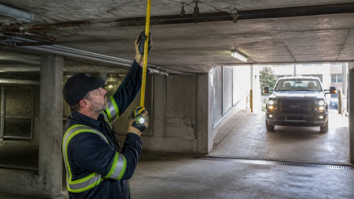 Professional tow truck operator measuring ceiling clearance in an underground parking garage with measuring tape and flashlight, examining the garage entrance carefully