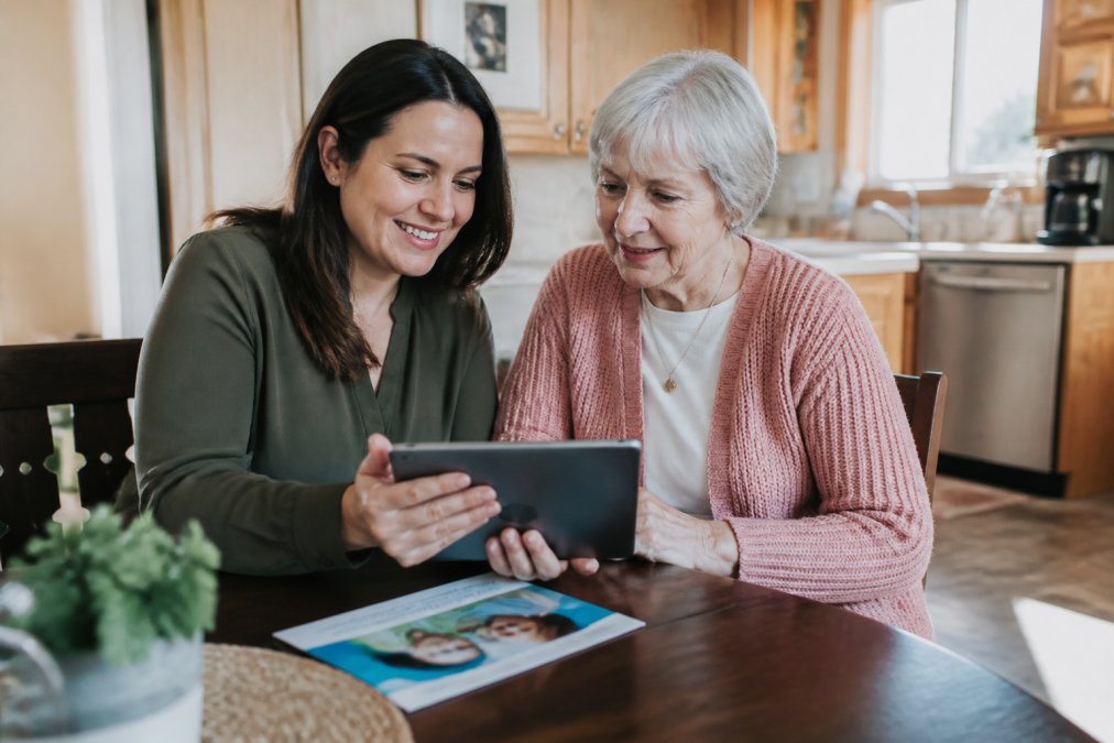 An adult daughter sitting at a kitchen table with her elderly mother, both looking at a tablet together with a caregiver brochure visible on the table