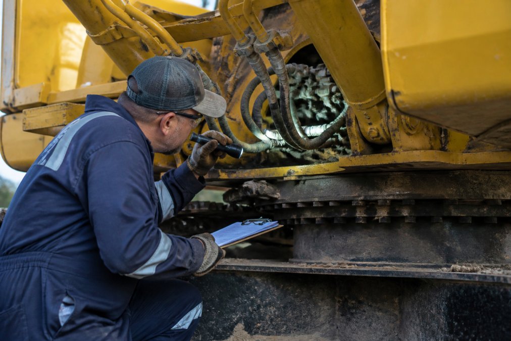 Technician in work clothes using a flashlight and clipboard to inspect the undercarriage and hydraulic systems of a yellow excavator, examining hoses and connectors closely