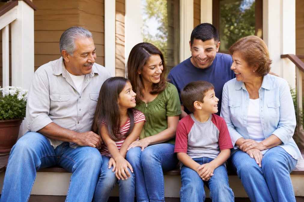 A multigenerational family gathered together outdoors on a sunny afternoon, including young children, parents, and grandparents sitting together on a porch, representing different life stages and the need for financial planning