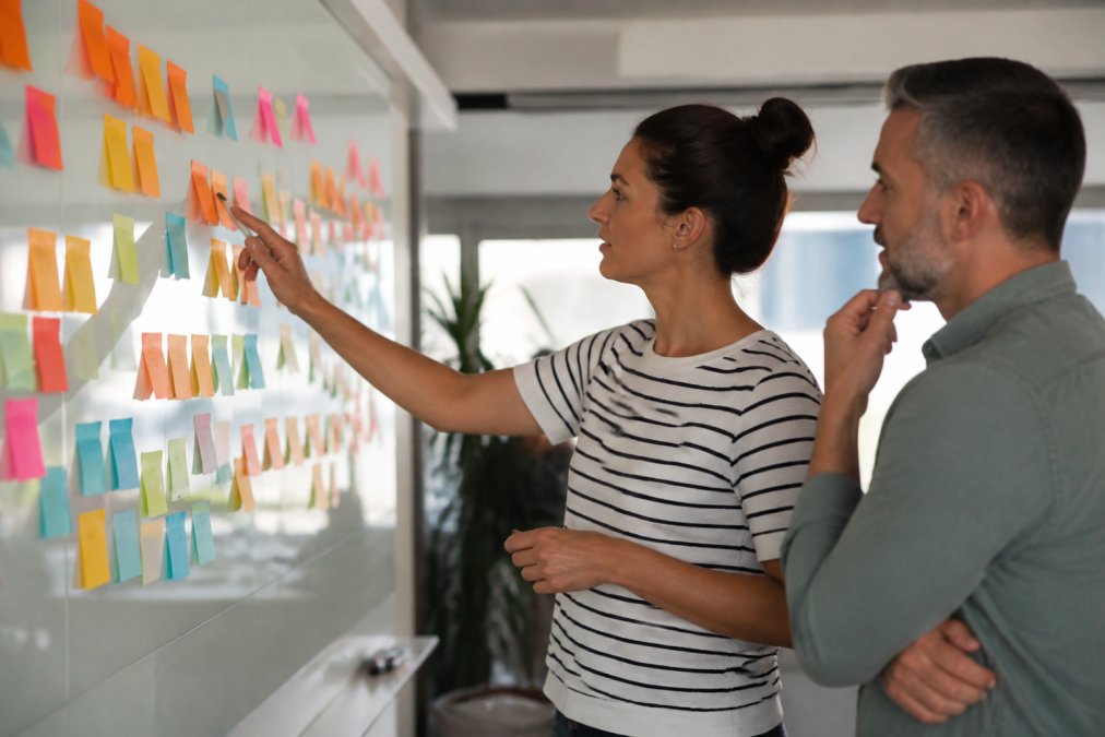 A Scrum Master and team lead standing at a wall covered in sticky notes, mapping out a transformation plan together in a bright meeting room