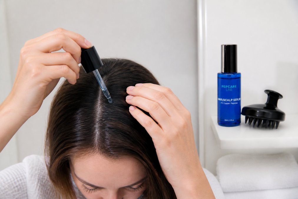 Hands applying a dropper serum directly onto a clean scalp, with a small scalp brush and serum bottle visible on a white bathroom shelf nearby, soft bathroom lighting