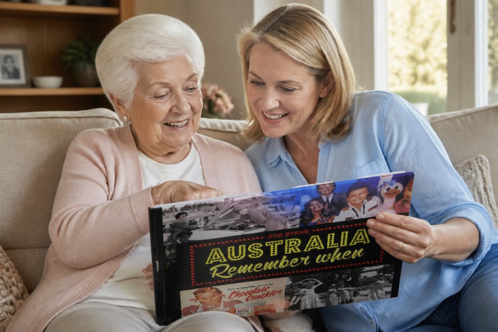 An elderly person and adult family member sitting together on a comfortable sofa, looking through a decade-specific photo book, both smiling and pointing at nostalgic images, warm home setting with natural afternoon light streaming through windows