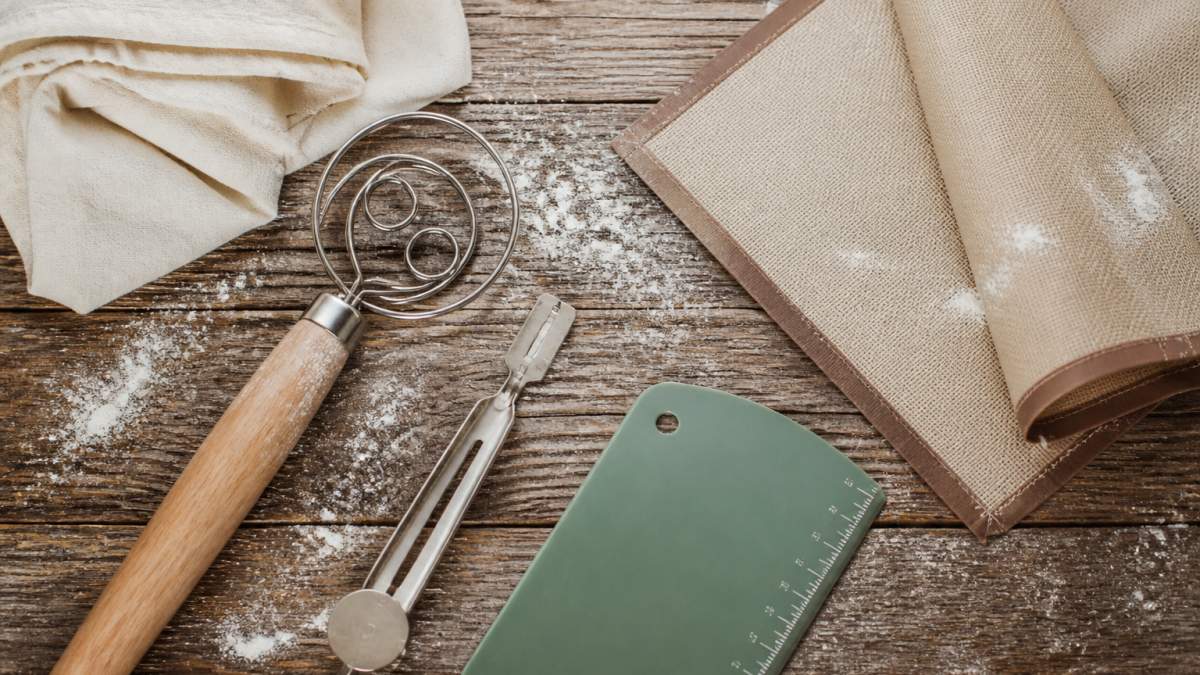 Flat lay of sourdough baking tools arranged on a weathered wooden surface: a stainless steel dough whisk with wooden handle, magnetic retractable bread lame with razor blades, green stainless steel dough scraper with measurement markings, silicone bread sling, and cream-colored cotton proofing cloth, with flour dusted artfully around the items