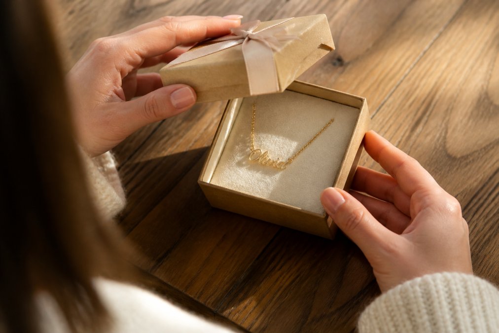 Close-up of hands opening a small gift box to reveal a personalized name necklace on a soft cloth, warm natural light on a wooden table