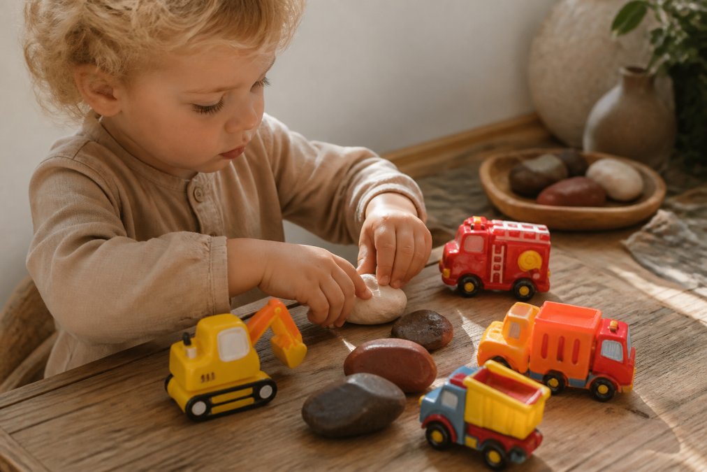 Een klein kind met blonde krullen dat voorzichtig speelt met natuurlijke speelklei in verschillende warme kleuren (terra, crème, bruin), kleine handen kneden zachtjes, op een houten tafel met daglicht van opzij
