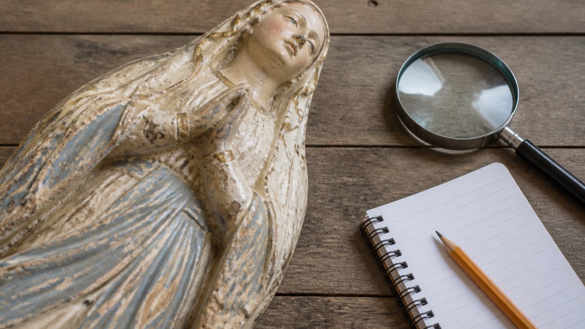 Primo piano di un'antica statua sacra in gesso con patina visibile e segni del tempo, accanto a una lente d'ingrandimento e a un taccuino su un tavolo di legno
