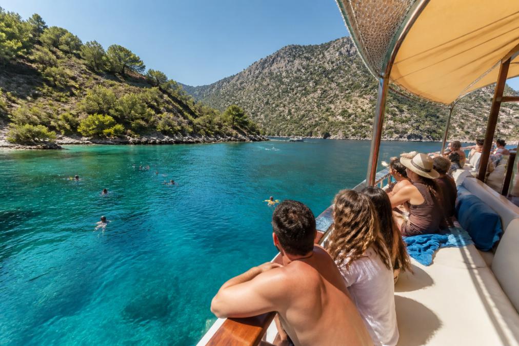 Güneşli bir günde Fethiye'de tekne turundan görüntü, denizde yüzen insanlar ve arka planda dağlık kıyı, teknede oturan gezginler ve turkuaz su