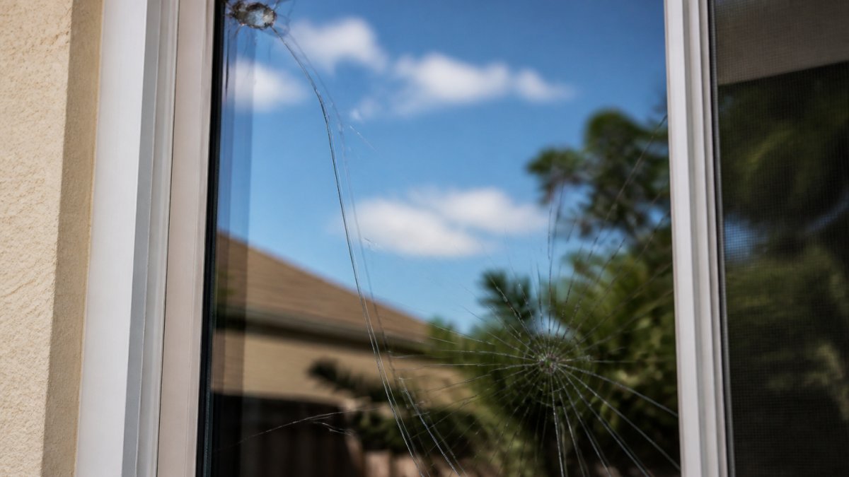 Close-up of a residential window pane showing various damage types: a small chip in the upper corner, a hairline crack running vertically, and a larger spiderweb fracture pattern in the lower section, with natural light revealing the damage clearly