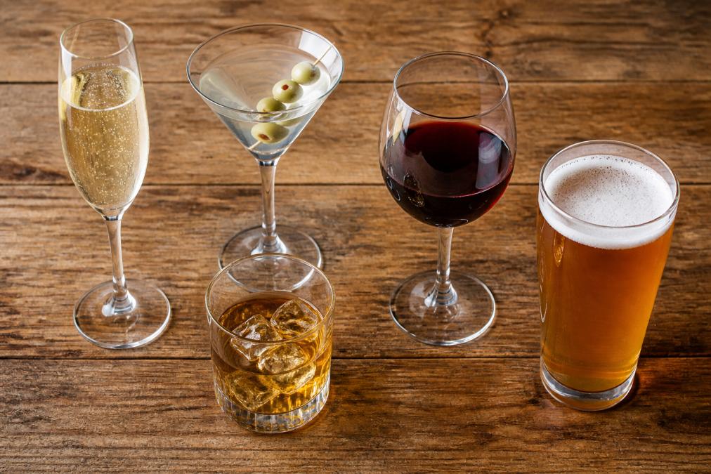 Close-up overhead shot of five different glass types arranged on a wooden table: champagne flute, martini glass, rocks glass, wine glass, and beer glass, each containing a different colored beverage, showing the distinct shapes and sizes