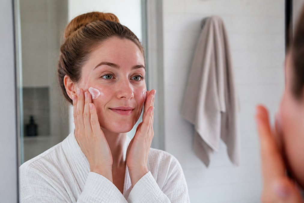 Woman with sensitive skin gently applying moisturizer to her face in natural bathroom lighting, showing proper application technique with upward circular motions