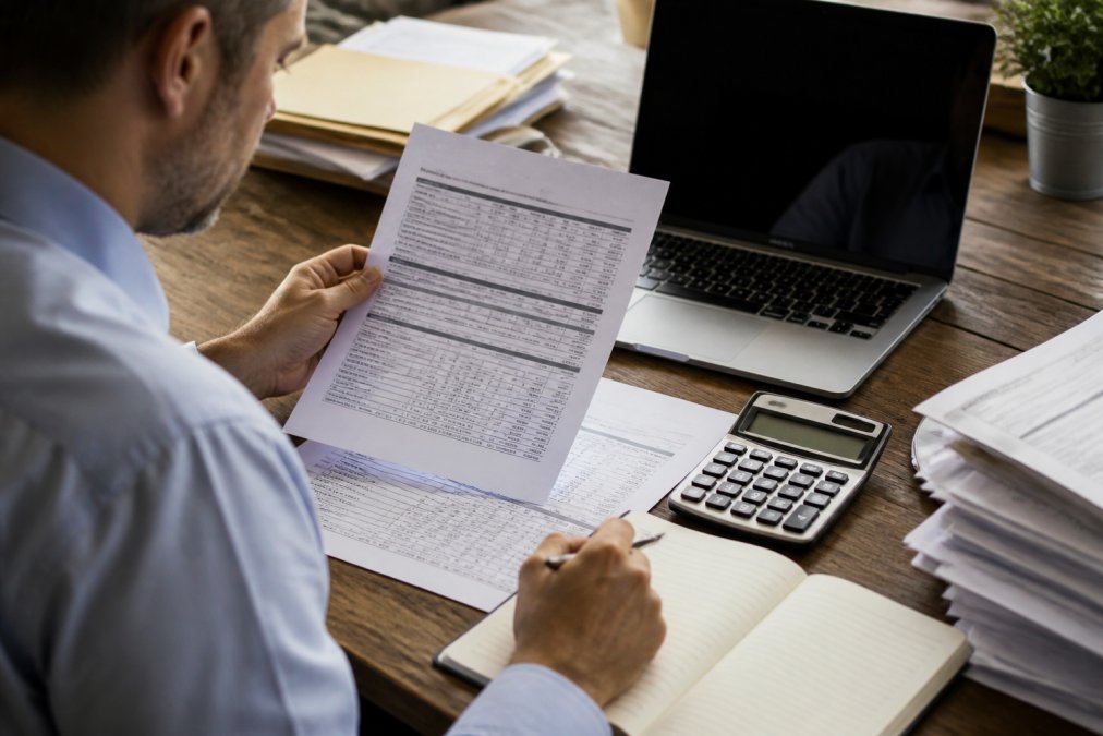Logistics manager reviewing detailed shipping invoices and freight cost documents at a desk with calculator, notepad, and laptop displaying rate analysis, natural office lighting