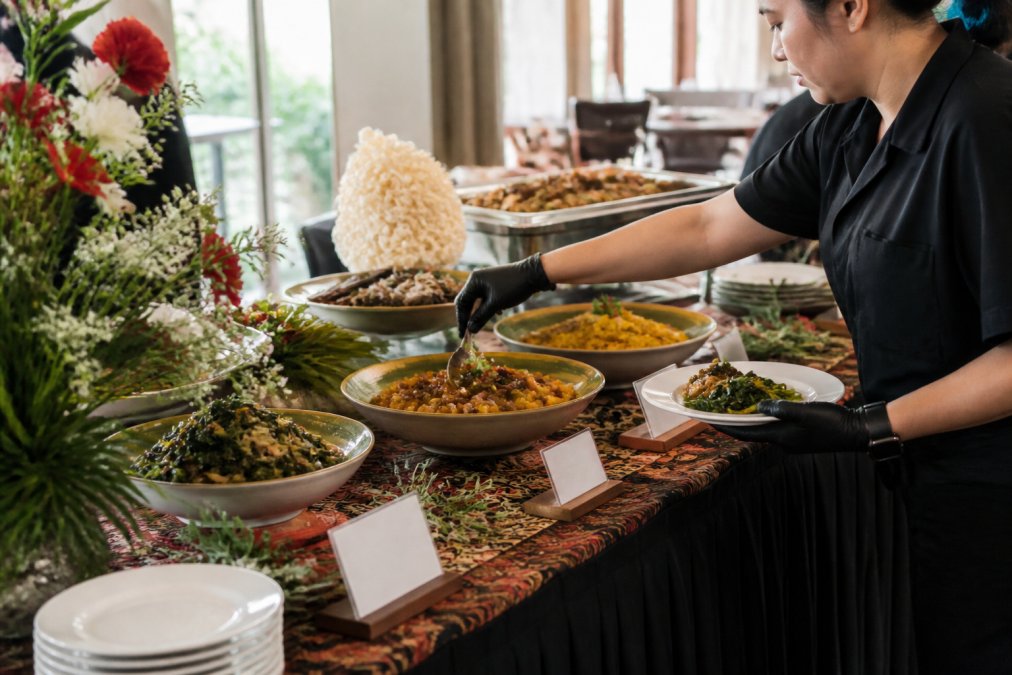A caterer in gloves carefully plating gluten free Indonesian dishes on a buffet table, with labelled dietary cards and fresh garnishes visible in a bright function room