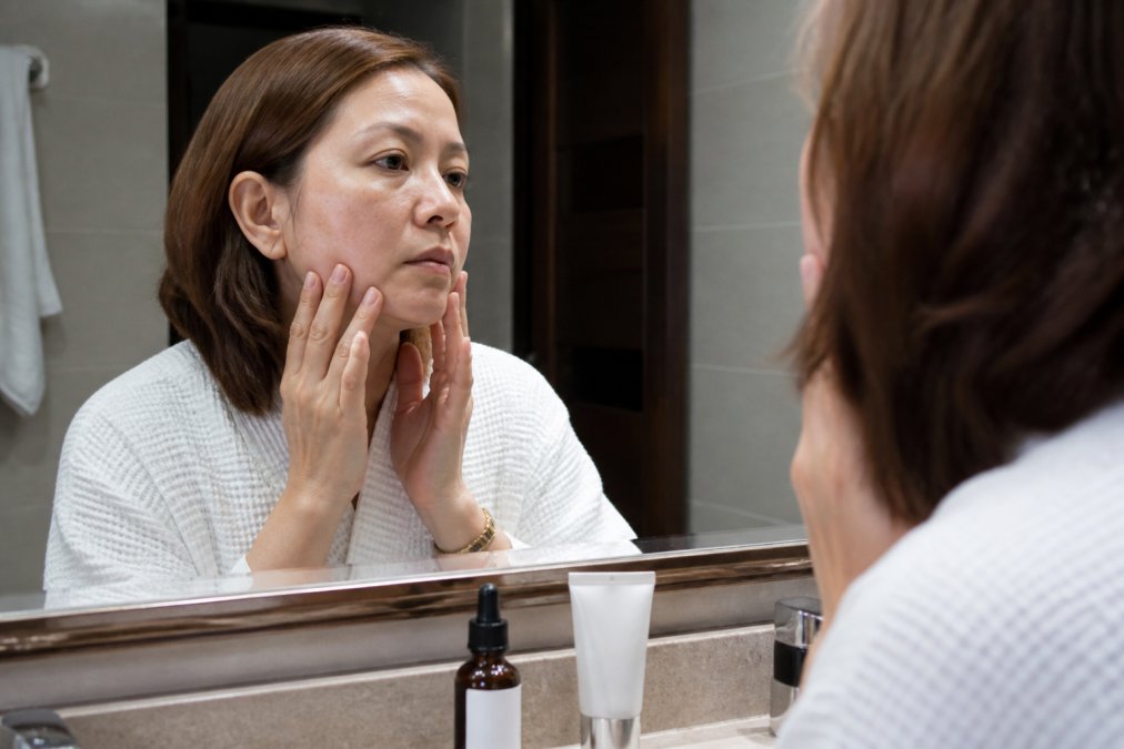 A woman in her 40s examining her skin in a bathroom mirror, with a bottle of glycolic acid peel and sunscreen on the counter