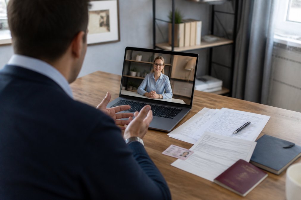 International entrepreneur on video call with bank representative, laptop screen displaying banking application forms and identity verification documents spread on desk in home office setting