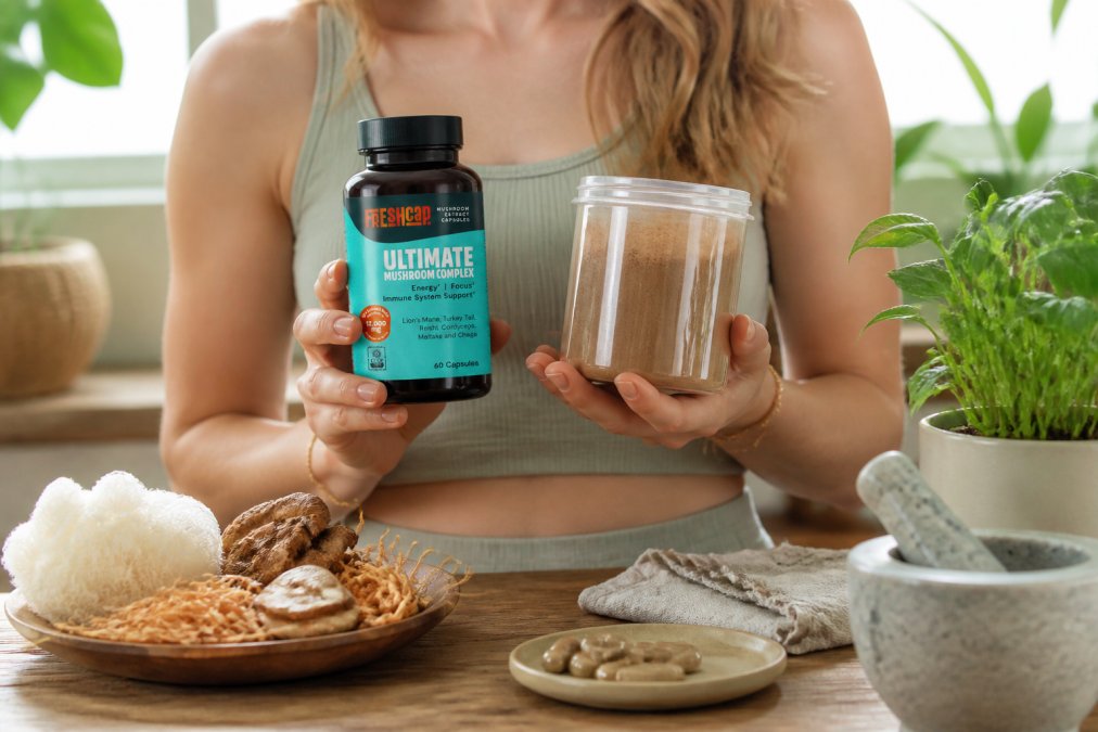 Person holding premium supplement capsules and powder containers in a bright, minimalist wellness space with natural lighting, displaying organic mushroom complex and herbal formulas