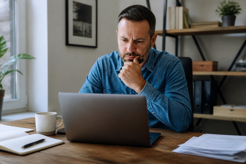 Small business owner sitting at desk reviewing website analytics on laptop, looking thoughtful with coffee cup nearby
