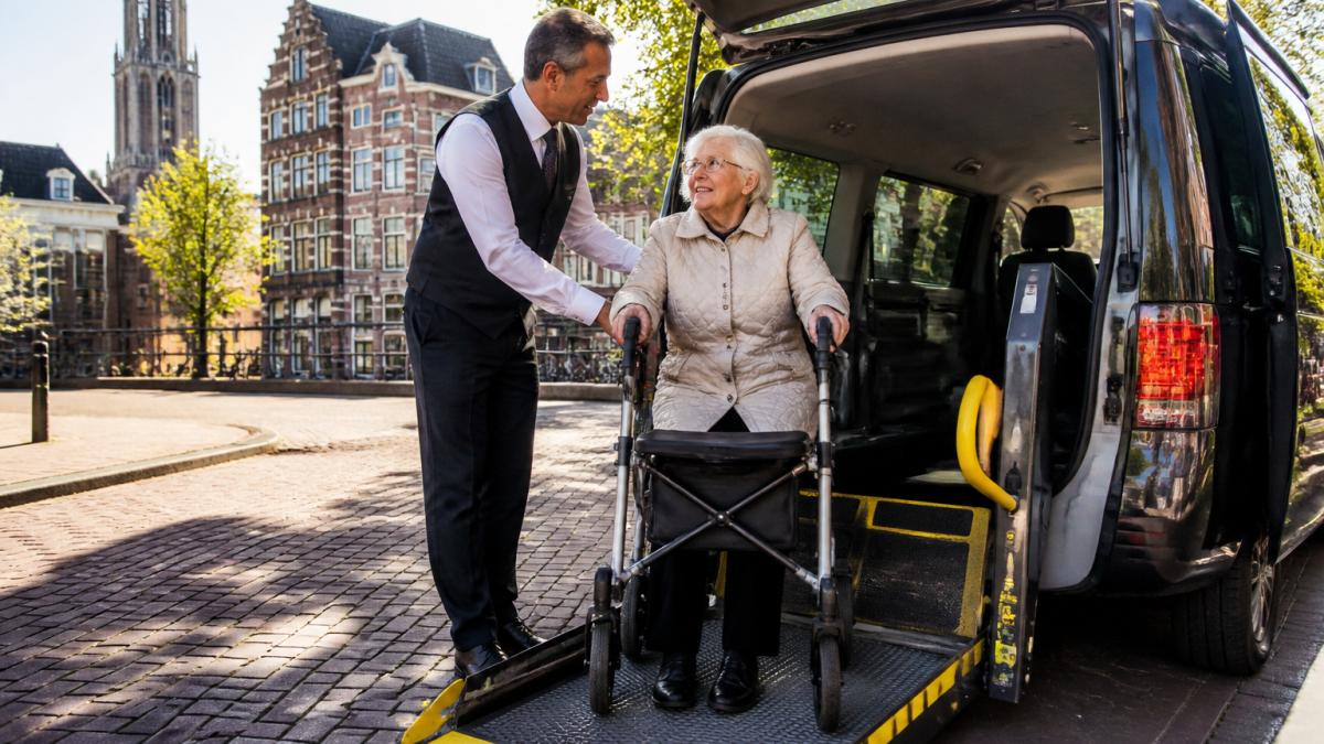 Een vriendelijke taxichauffeur in uniform helpt een oudere mevrouw met een rollator bij het instappen in een rolstoeltoegankelijke taxi op een zonnige straat in Utrecht