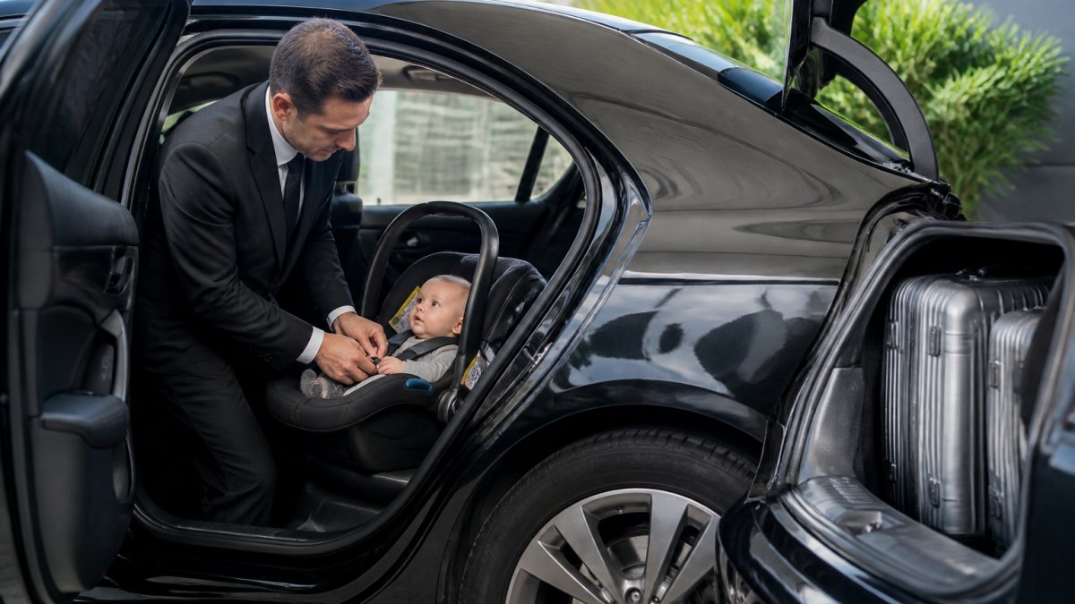 Professional chauffeur securing a rear-facing infant car seat in the back of a luxury sedan, with child visible in seat and luggage loaded in the boot, natural daylight through vehicle window