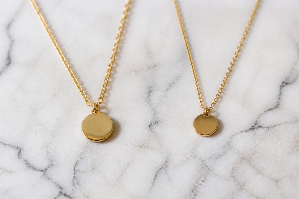 Close-up flat lay of two delicate gold necklaces side by side on a white marble surface, one visibly thicker and more lustrous than the other, with soft natural lighting highlighting the surface finish differences