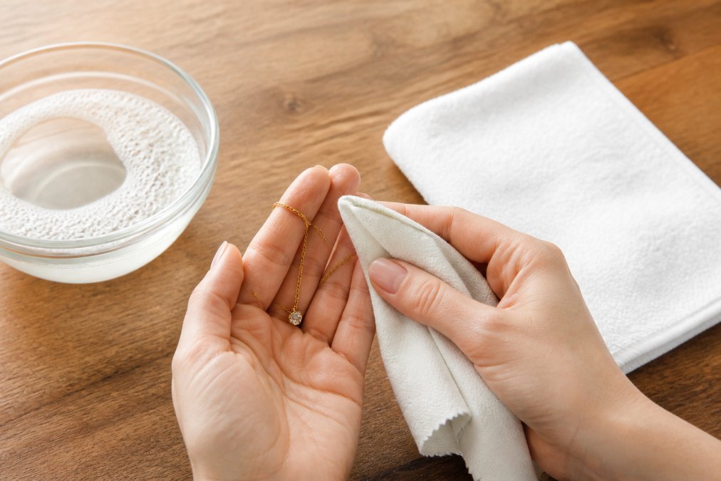 Hands gently wiping a delicate gold necklace with a soft microfiber cloth on a clean wooden surface, with a small bowl of warm soapy water and a dry towel nearby in soft natural light
