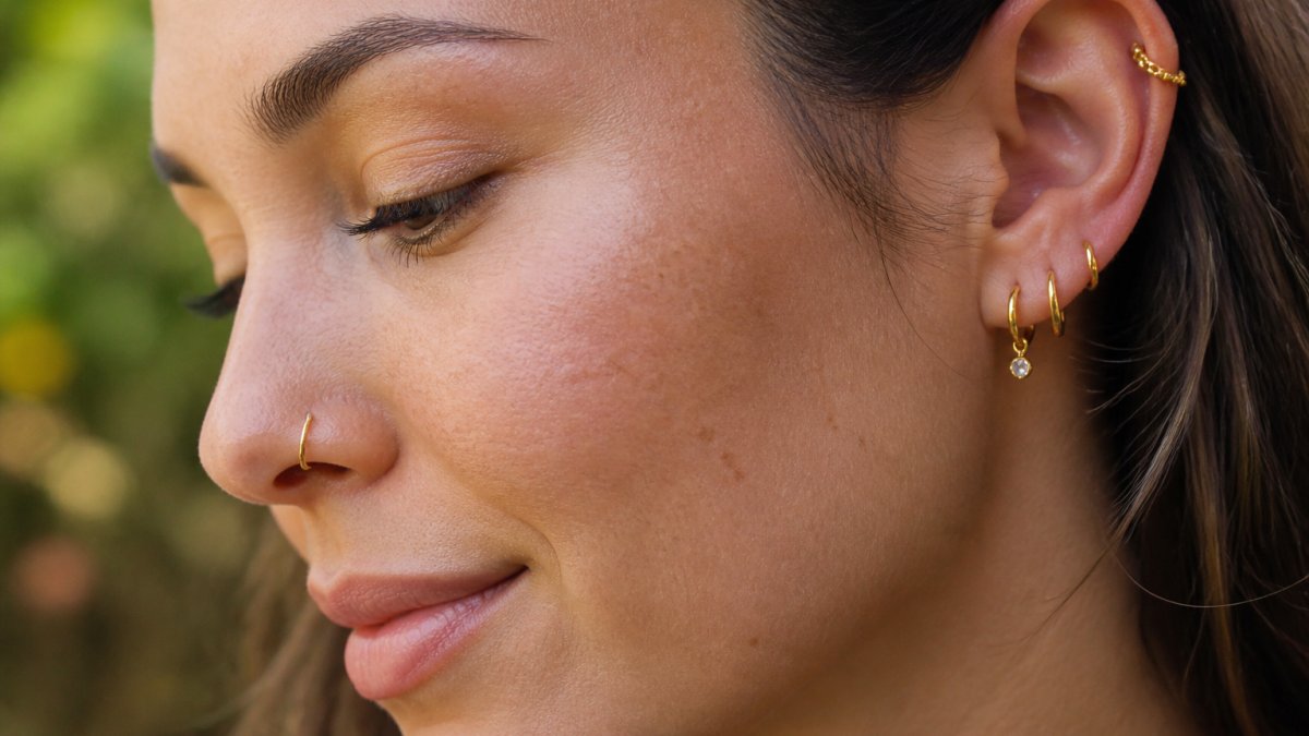 Close-up of a woman wearing delicate 14K gold piercing jewelry in her helix and nose, showing the jewelry detail and skin tone against natural daylight