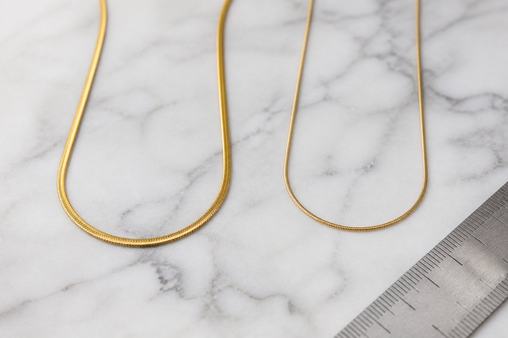 Close-up flat lay of two gold necklaces side by side on a white marble surface, one with a warm, thick gold finish and one with a thinner, slightly duller finish, with a small ruler nearby suggesting measurement