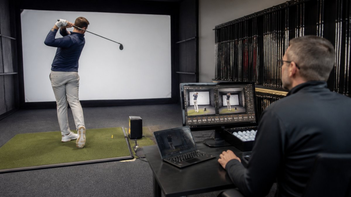 Professional PGA instructor using launch monitor technology to analyse golfer's swing in private fitting studio, with golfer mid-swing and technology display visible showing real-time data