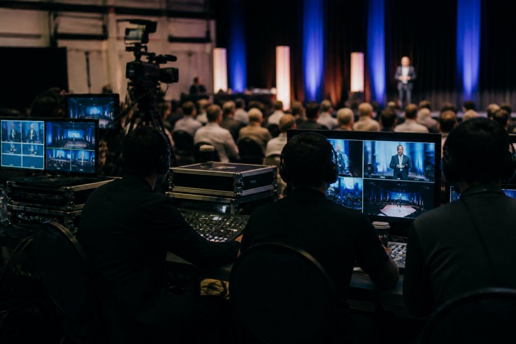 Professional event production team managing multiple camera feeds and streaming equipment at a control station during a live corporate presentation, with monitors displaying real-time analytics and attendee engagement metrics