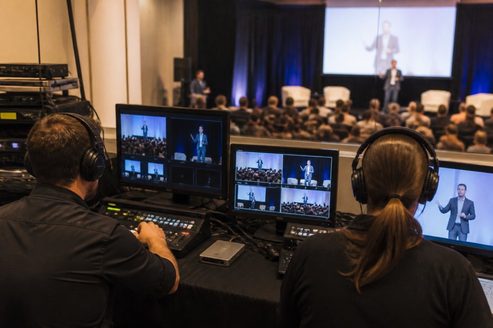 Event production control room with operators monitoring multiple screens showing live feed from conference stage, remote attendee engagement metrics, and technical performance indicators during a large corporate event