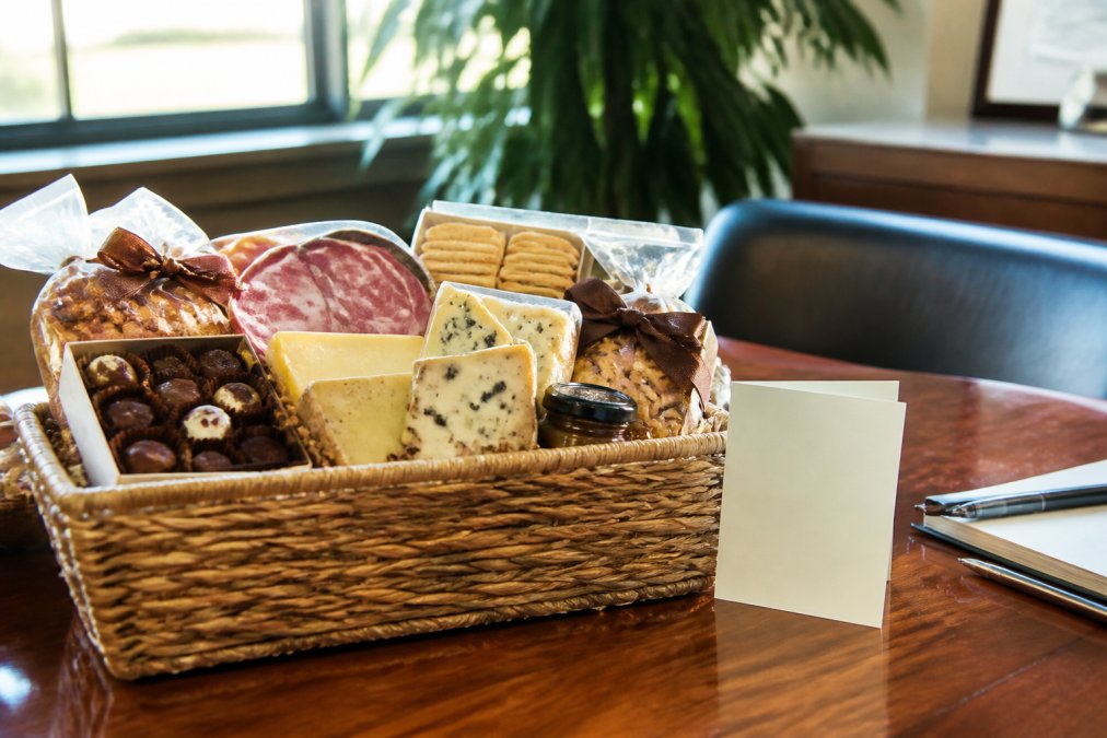 A beautifully arranged gourmet gift basket on a wooden desk in a bright office, featuring artisan cheeses, cured meats, crackers, and chocolates, with a handwritten note card tucked beside it