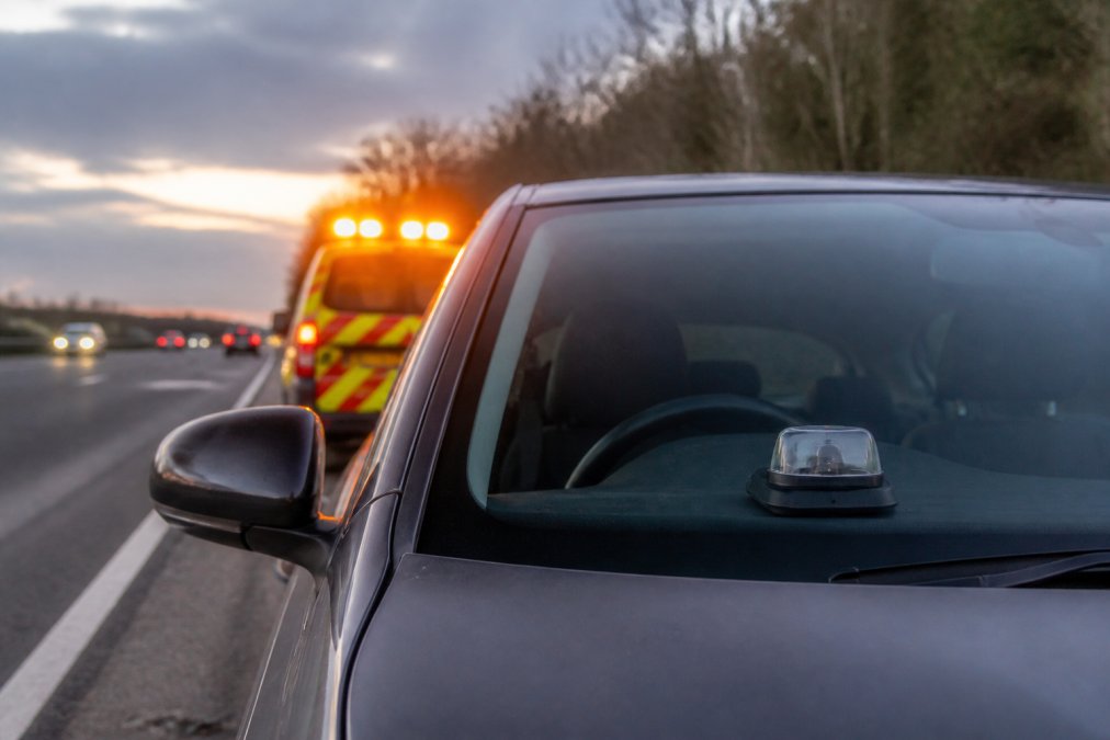 GPS Beacons for Safety: Benefits & How They Work A stationary vehicle on a motorway hard shoulder at dusk with high-visibility warning lights activated and a GPS beacon mounted on the dashboard, showing emergency hazard awareness in action