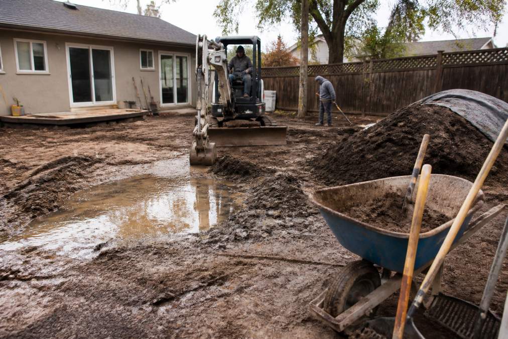 Landscaping crew using a compact excavator to regrade muddy, uneven yards with fresh topsoil