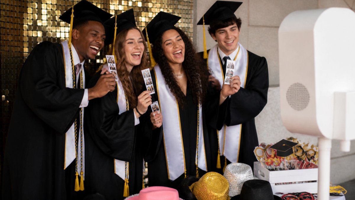 A group of graduates in caps and gowns posing together in front of a photo booth backdrop, laughing and holding printed photo strips, with a prop table of graduation-themed hats and signs nearby