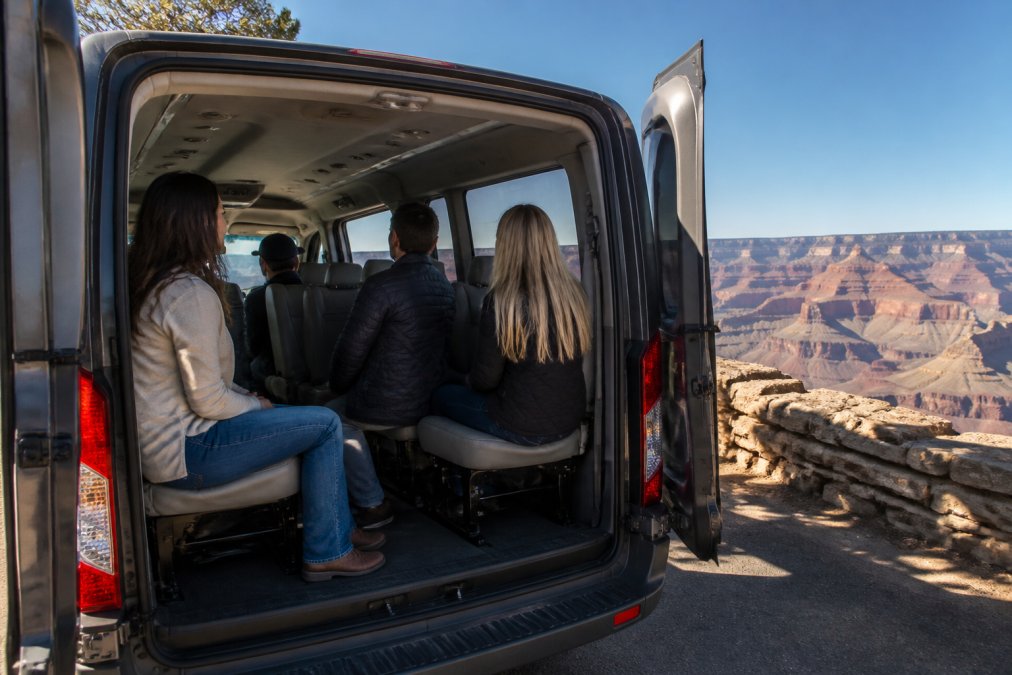 A Ford Transit van with open rear doors parked at a scenic overlook, showing comfortable interior seating with four passengers gazing at the expansive Grand Canyon vista in the background, natural daylight streaming through large windows