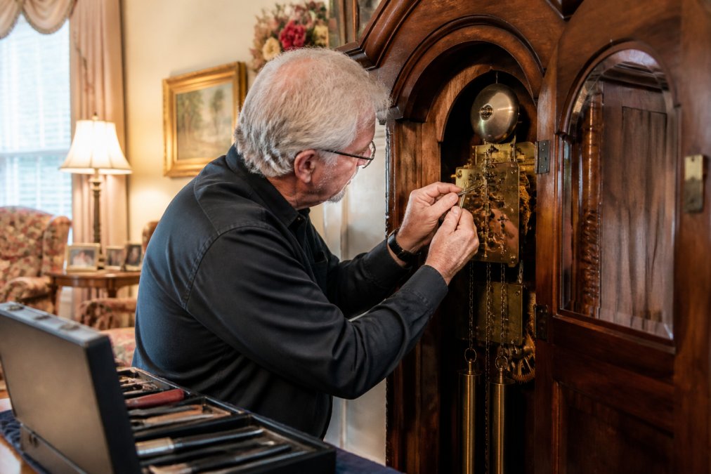 Professional clockmaker examining a movement during an in-home grandfather clock repair service in a warm living room
