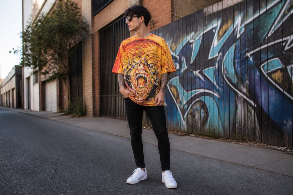 Person wearing an oversized graphic streetwear t-shirt in bold saturated colors, styled with fitted black jeans and clean white sneakers, standing in natural daylight on an urban street showing street-ready aesthetic
