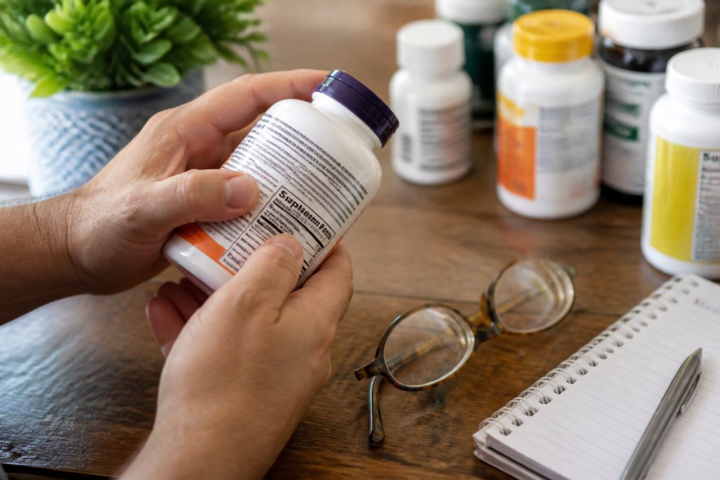 Close-up of hands holding a dietary supplement bottle, examining the label and ingredient list in natural daylight with reading glasses nearby