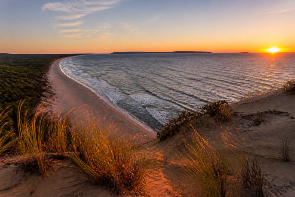 Aerial view of the Sleeping Bear Dunes at sunset, with the Manitou Islands visible on the horizon and waves crashing on the sandy shore